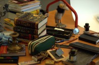 Chaos - Auf einem Tisch liegen Stapel Bücher, ein Kerzenständer, eine Raben-Figur, Stifte, ein Mäppchen eine alte Schiefertafel und Schieferstifte verstreut.
