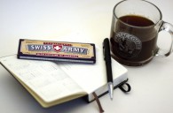 Caffeine - Auf einem Notizbuch liegt eine Tafel "Swiss Army" Schokolade mit Koffein und ein Kugelschreiber. Daneben steht eine durchsichtige Tasse die Kaffee enthält.