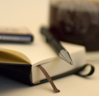 Write - Ein Kugelschreiber liegt auf einem Notizbuch, dahinter steht ein Glas Kaffee. Nur die Spitze des Kugelschreibers ist im Fokus.